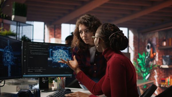 One woman gesturing towards her computer while conversing about coding with another woman