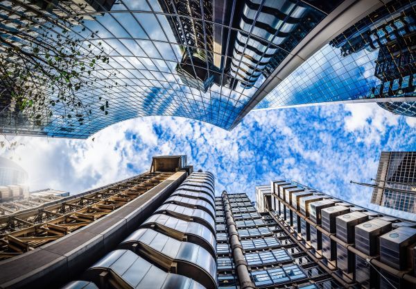 An up shot of buildings up towards the sky during slightly cloudy weather. The buildings are extremely reflective and reflect the clouds from the sky